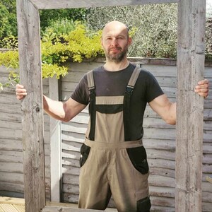 May include: A man wearing brown overalls stands in front of a large, weathered wooden frame. The frame is held up by the man's hands. The frame is in front of a wooden fence and green foliage.