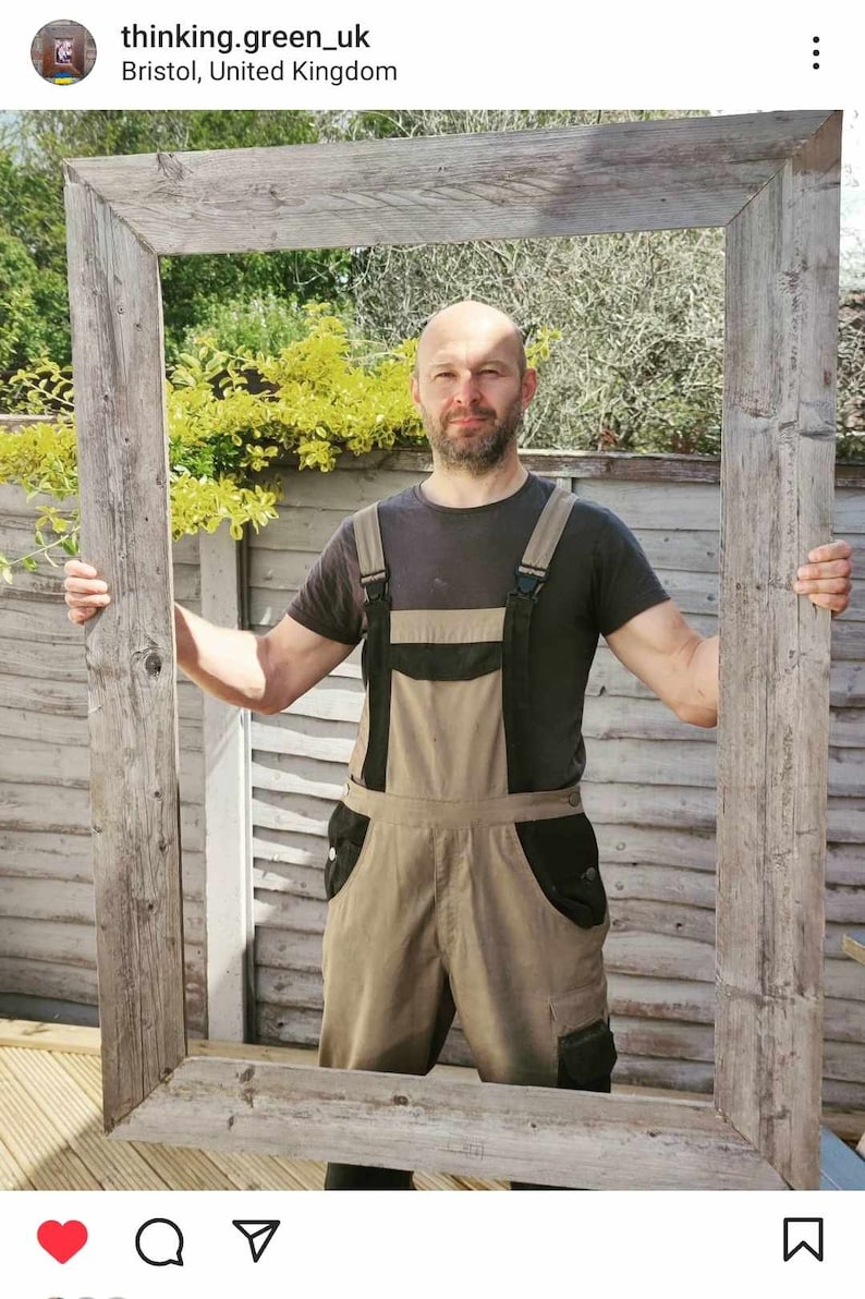 May include: A man wearing brown overalls stands in front of a large, weathered wooden frame. The frame is held up by the man's hands. The frame is in front of a wooden fence and green foliage.