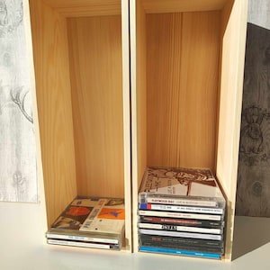 May include: Two wooden shelves with a variety of music CDs. The shelves are made of light wood and are empty except for the CDs. The CDs are all different colours and have different artists and album titles. The CDs on the right shelf include Fleetwood Mac, Sting, and The Rolling Stones.