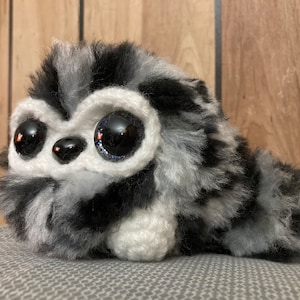 Könnte beinhalten: Ein Plüsch-Waschbär-Spielzeug mit schwarz-weißem Fell, großen schwarzen Augen und einer weißen Nase. Das Spielzeug liegt auf einer grauen Stoffoberfläche.