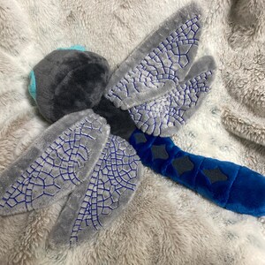 May include: A plush dragonfly toy featuring a dark gray fuzzy head with bright blue eyes, four gray wings with intricate blue embroidered patterns, and a long, segmented blue body decorated with dark gray diamond-shaped accents.