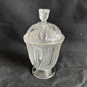 May include: Clear glass candy dish with a lid. The dish has a detailed, textured pattern covering the entire surface, including the lid and the base. The lid has a decorative handle. The dish is elevated on a small, round base.