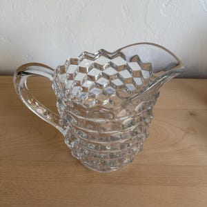 May include: A clear glass pitcher with a textured, diamond-patterned exterior. The pitcher has a handle and a spout for pouring. The design is repeated around the body of the pitcher. The pitcher is sitting on a wooden surface.