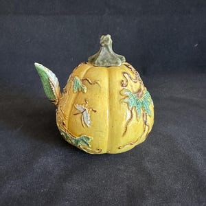 May include: A yellow ceramic teapot shaped like a pumpkin with a green stem lid. The surface features raised relief details including green leaves, brown vines, and a small white insect. A green leaf-shaped spout extends from the side of the body.