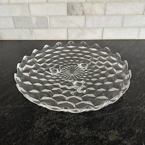 May include: A clear glass cake stand featuring a textured diamond pattern across the surface, a scalloped edge, and three small feet. The center of the plate displays a radial sunburst design, and the overall construction is transparent glass.