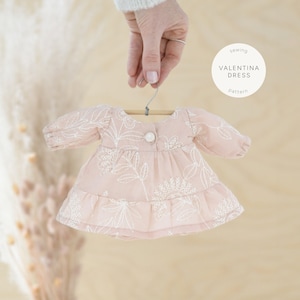 Peut inclure: Une robe rose avec des broderies florales blanches, suspendue à un cintre en bois. La robe a des manches longues et une jupe plissée. Le texte "couture Valentina Dress Pattern" est sur un cercle blanc.