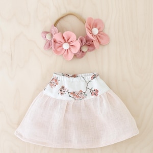 Peut inclure: Un bandeau orné de fleurs en feutrine dans des tons rose et beige avec des centres blancs, accompagné d'une jupe dotée d'une ceinture blanche imprimée d'un motif floral de cerisier et d'une partie inférieure froncée rose pâle.