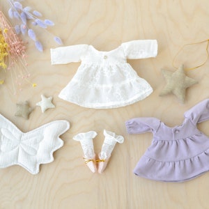 May include: A set of doll clothes including a white lace dress with long sleeves, a purple dress with long sleeves, a pair of white lace socks, and a pair of white wings.
