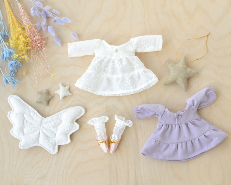 May include: A set of doll clothes including a white lace dress with long sleeves, a purple dress with long sleeves, a pair of white lace socks, and a pair of white wings.