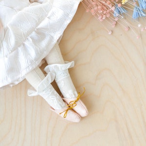May include: A white doll with a white lace dress and pink shoes with gold bows. The doll is lying on a light wood surface with dried flowers in the background.