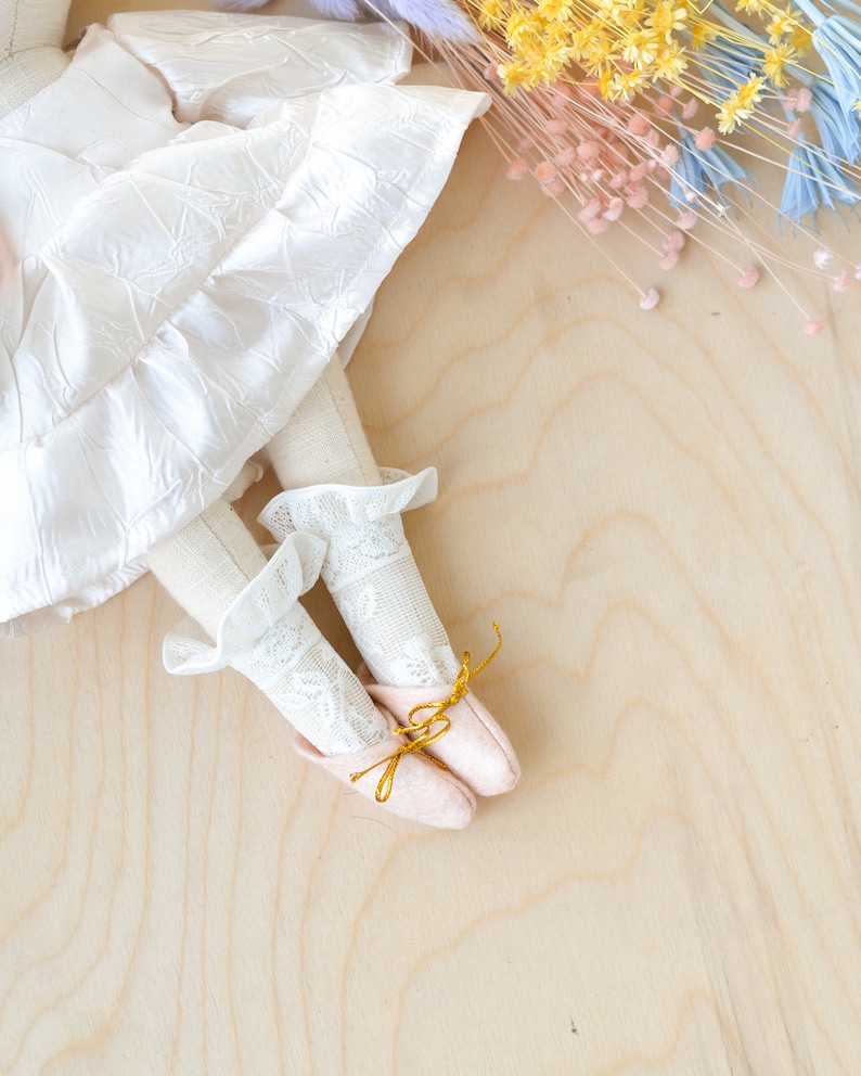 May include: A white doll with a white lace dress and pink shoes with gold bows. The doll is lying on a light wood surface with dried flowers in the background.