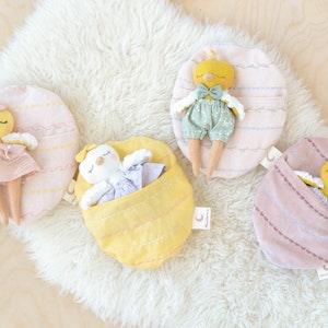 Peut inclure: Quatre poupées poulets jaunes et blanches dans différentes poses, chacune allongée dans un nid en tissu rayé rose ou jaune. Les nids ont une poche sur le côté et une petite étiquette avec les mots "Sleepytime Crew" dessus.