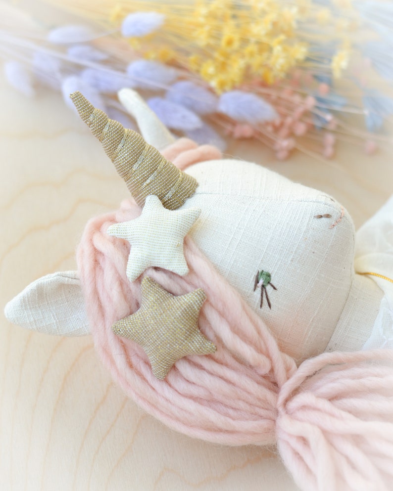 May include: A white unicorn doll with a gold horn and pink yarn hair. The doll has two gold glitter stars and one white star sewn on its head.