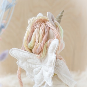 May include: A white fabric unicorn doll with a gold horn and pink and yellow yarn hair. The doll is wearing a white dress with a floral pattern and has white wings.