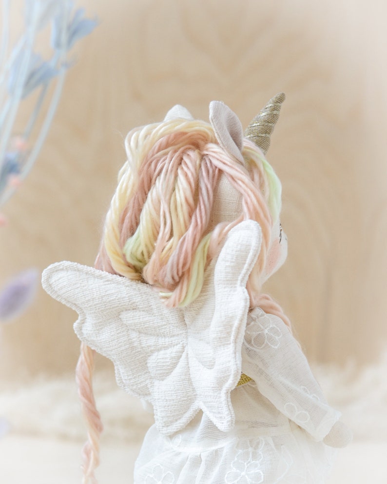 May include: A white fabric unicorn doll with a gold horn and pink and yellow yarn hair. The doll is wearing a white dress with a floral pattern and has white wings.