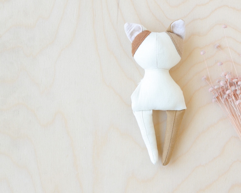 K&ouml;nnte beinhalten: Eine wei&szlig;e und braune Stoffkatzenpuppe mit schlichtem Design. Die Puppe liegt auf einer hellen Holzoberfl&auml;che.