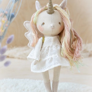 May include: A handmade white unicorn doll with a pink and yellow yarn mane and tail. The doll is wearing a white dress with embroidered flowers and has a gold horn.