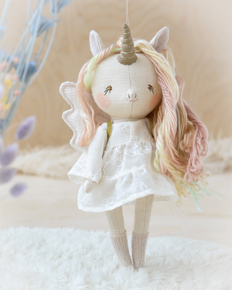 May include: A handmade white unicorn doll with a pink and yellow yarn mane and tail. The doll is wearing a white dress with embroidered flowers and has a gold horn.