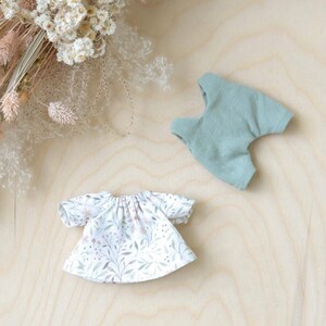 K&ouml;nnte beinhalten: Zwei Puppenoutfits, eines ein wei&szlig;es Kleid mit Blumenmuster und das andere eine Salopette in Salbeigr&uuml;n.
