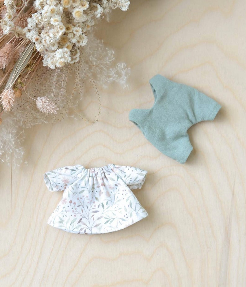 K&ouml;nnte beinhalten: Zwei Puppenoutfits, eines ein wei&szlig;es Kleid mit Blumenmuster und das andere eine Salopette in Salbeigr&uuml;n.