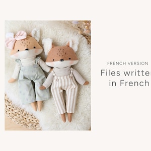 May include: Two plush fox toys with brown fur, white accents, and black eyes. One fox is wearing a light blue overall with white floral print and a pink bow. The other fox is wearing a light brown overall with white stripes. The toys are sitting on a white fluffy surface.  French Version: Files written in French.
