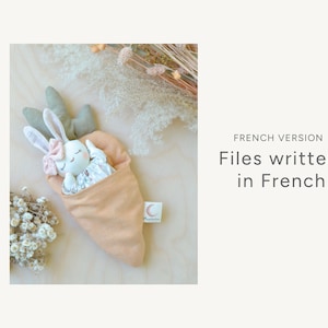 May include: A white bunny doll with pink accents is sleeping inside a peach-coloured carrot-shaped fabric pouch. The pouch has a small tag with a crescent moon and the words "Magpie Lane".