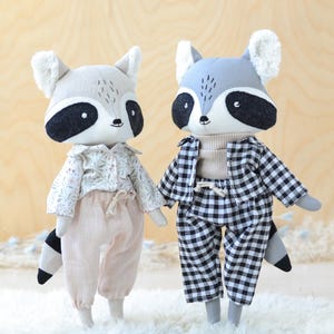 Peut inclure: Deux jouets en peluche en forme de raton laveur portant des tenues différentes. Un raton laveur porte une chemise à imprimé floral et un pantalon beige. L'autre raton laveur porte une chemise et un pantalon à carreaux noirs et blancs.