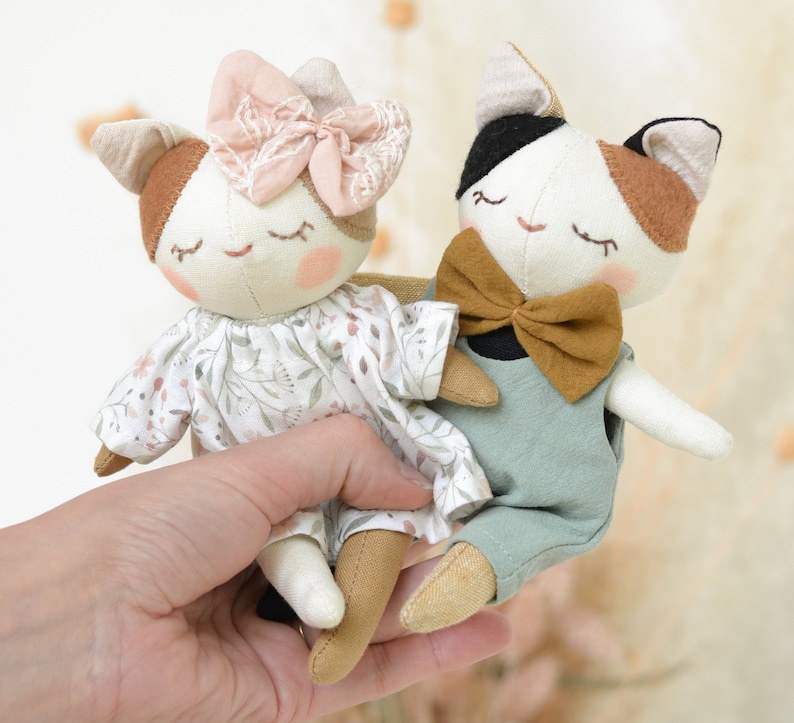 K&ouml;nnte beinhalten: Zwei handgefertigte Pl&uuml;schkatzenspielzeuge. Eine Katze tr&auml;gt ein Blumenkleid mit einer rosa Schleife. Die andere Katze tr&auml;gt eine blaue Latzhose mit einer braunen Fliege.