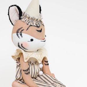 May include: A fabric tiger doll wearing a cream-colored cone hat with a ruffled collar, a matching cream neck ruff, and tan and black striped trousers with two small white pom-poms. The doll features embroidered black stripes and pink cheeks.