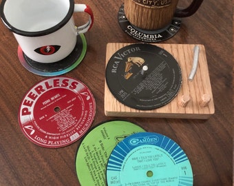 Puede incluir: Cuatro posavasos de vinilo de diferentes colores: rojo, azul, verde y negro. Los posavasos muestran el texto "Peerless", "RCA Victor", "Camden" y "Columbia Masterworks".