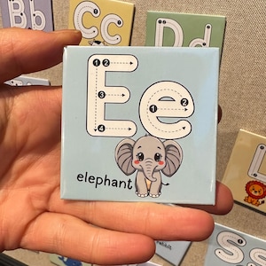 May include: A square educational card featuring the uppercase and lowercase letter E in white with black directional arrows and numbers for stroke order. Below the letter is a cartoon illustration of a grey elephant and the word elephant in black.