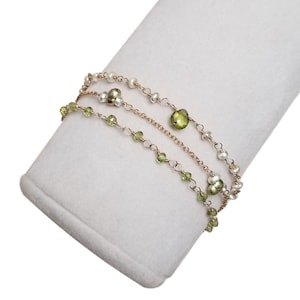 May include: A three-strand bracelet featuring gold chains, small white pearls, and green peridot gemstones. The bracelet is displayed on a white, angled surface, showcasing its delicate design and color contrast.