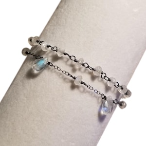 May include: A delicate bracelet with two strands of small white beads and teardrop-shaped charms. The bracelet is made with black wire and silver chain.