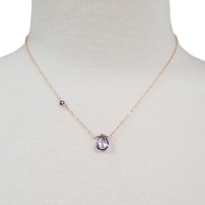 May include: A delicate gold chain necklace with a single teardrop-shaped amethyst gemstone pendant. The necklace features a small black bead accent.