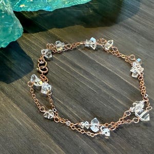 May include: A delicate rose gold bracelet featuring clear crystal accents. The double-strand chain is adorned with irregularly shaped crystals, creating a sparkling and elegant accessory. The bracelet is displayed on a dark wooden surface.