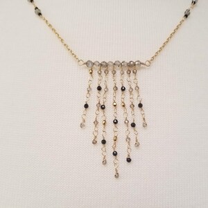 May include: A gold chain necklace with a cascading fringe of black and gray beads. The necklace features a row of gray beads at the top, with strands of beads hanging down from it.