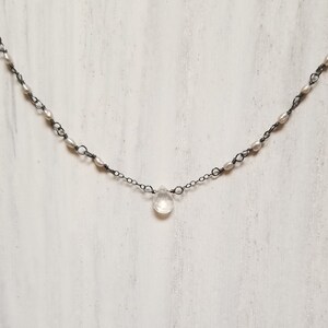 May include: A delicate necklace featuring a dark metal chain adorned with small, white pearls. A clear, teardrop-shaped pendant hangs from the center. The necklace is displayed against a white, textured background.