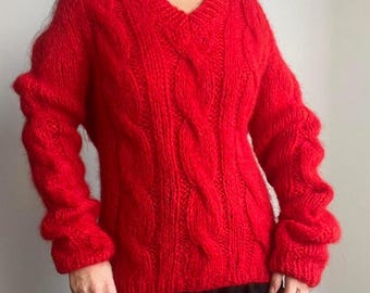 Op de afbeelding: Een felrode kabelgebreide trui met een V-hals. De trui heeft een textuurpatroon en lange mouwen. De heldere rode kleur en de dikke brei suggereren een warm en comfortabel kledingstuk, perfect voor koeler weer.