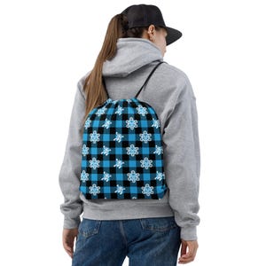 May include: A blue and black plaid drawstring backpack with a repeating pattern of white cartoon turtles. The backpack is being worn by a person wearing a gray hoodie, blue jeans, and a black baseball cap.