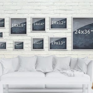May include: A white brick wall with a white couch and nine white picture frames with black and white images and measurements in inches. The frames are arranged in a staggered pattern.