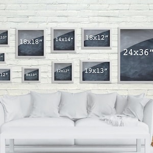 May include: A white brick wall with a variety of white picture frames of different sizes. The frames are all empty and have black text inside indicating the dimensions of each frame in inches. The frames are arranged in a staggered pattern on the wall.