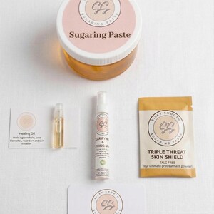 May include: A collection of skincare products, including a jar of Sugaring Paste, a bottle of Clarifying & Soothing Spray, a packet of Triple Threat Skin Shield, and a vial of Healing Oil. The products are labeled with the Silky Smooth Sugaring logo.