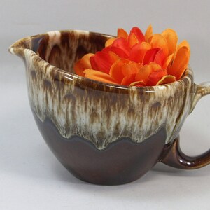 May include: A ceramic creamer pitcher featuring a glossy dark brown base and a cream-colored drip glaze pattern around the rim. The pitcher has a curved handle and a pouring spout, and it is filled with several bright orange and red artificial flowers.