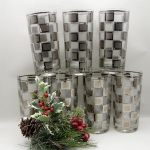 May include: A set of six clear glass tumblers featuring a repeating silver square checkerboard pattern and a textured finish. The glasses are arranged in two rows, accompanied by a decorative sprig of pine needles, holly leaves, and red berries.