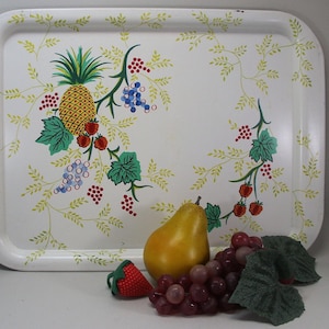 May include: A white rectangular serving tray with rounded corners, decorated with a pineapple, grapes, and strawberries. The fruit is painted in vibrant colors, with green leaves and yellow vines. A pear, grapes, and a strawberry are in front of the tray.