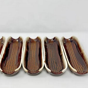 May include: A set of four elongated ceramic corn-shaped dishes featuring a deep brown glazed interior with a textured kernel pattern and a contrasting cream-colored rim. Each dish has a curved, trough-like form designed for serving corn on the cob.