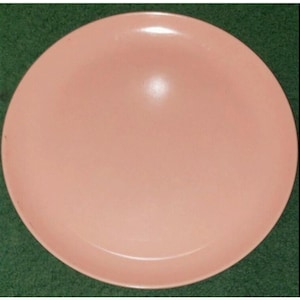 May include: A round, light pink plate with a smooth surface. The plate is a solid color and appears to be made of ceramic or a similar material. The plate is set against a green background.