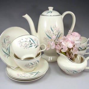 May include: A cream-colored ceramic tea set featuring a teapot, creamer, and multiple cups and saucers. Each piece is decorated with a delicate botanical pattern of thin teal leaves and small pink flowers, accented by a thin silver rim on the edges.