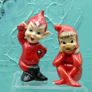 May include: Two ceramic elf figurines in red outfits. The left elf stands with a pointed hat, black boots, and a heart on its chest. The right elf sits with a red cap, brown hair, and hands near its face. Both have painted facial features.