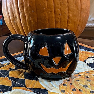 May include: A glossy black ceramic mug shaped like a jack-o'-lantern with a curved handle. The mug features recessed orange cutouts forming two triangular eyes and a wide, jagged, smiling mouth, creating a classic Halloween-themed drinkware piece.